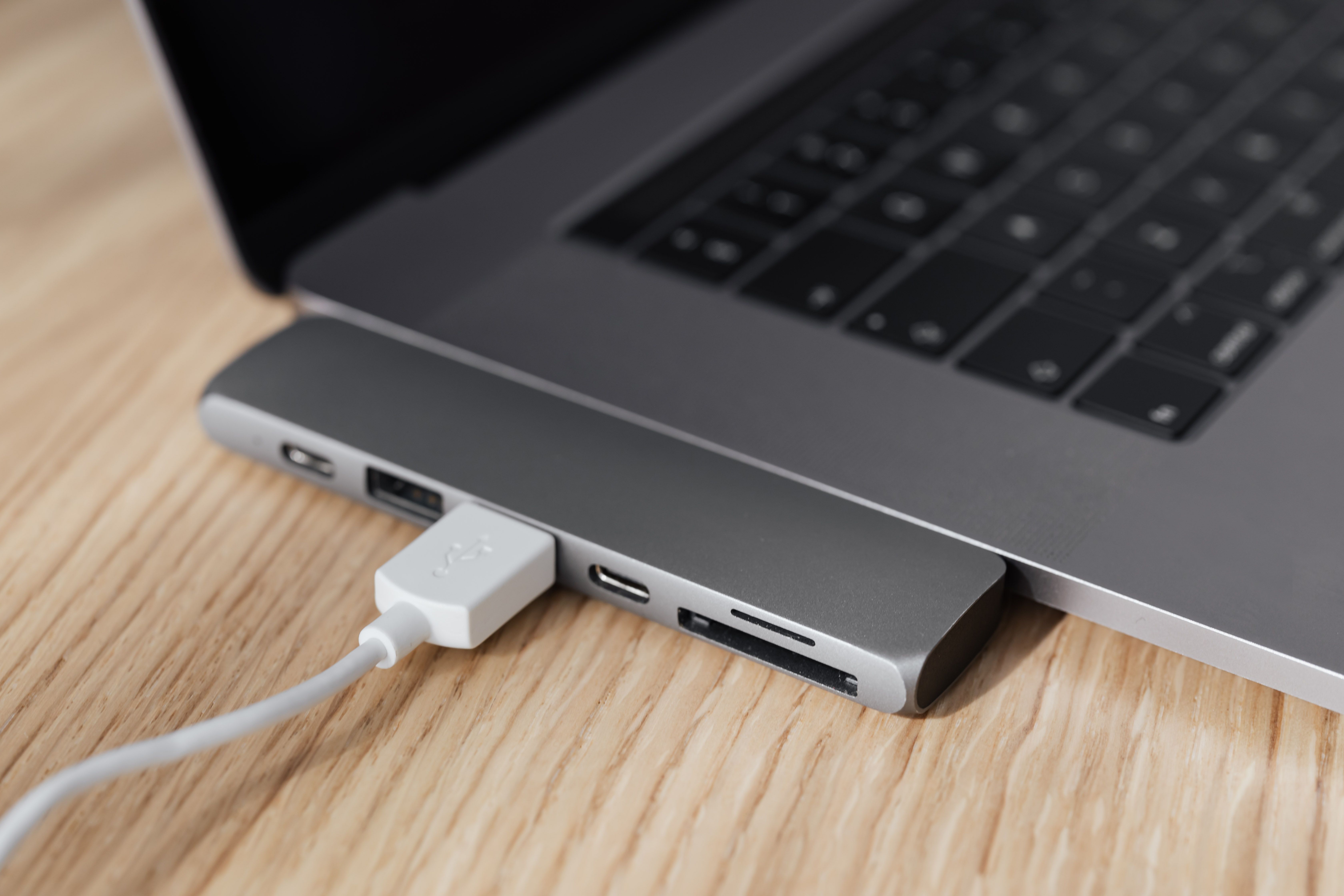 A USB-C hub plugged into the side of a silver laptop on a wooden desk, with a USB cable connected to one of its ports