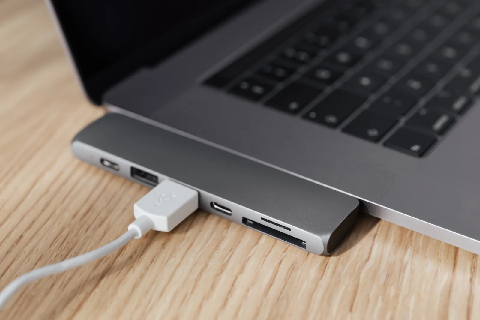 A USB-C hub plugged into the side of a silver laptop on a wooden desk, with a USB cable connected to one of its ports